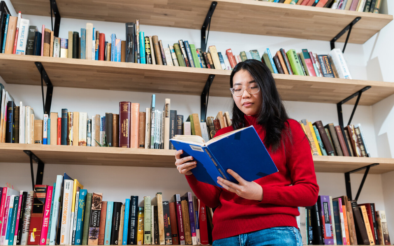 Image shows a student in a red knit jumper reading a book standing against a bookshelf