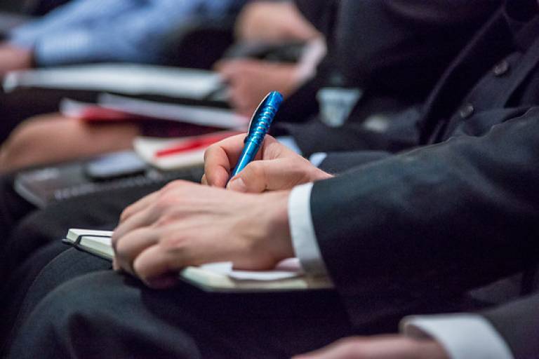 Row of people seated, one hand holding a pen by a notebook