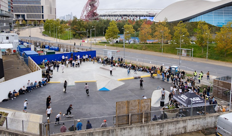 A wide shot from a distance of groups of people on a skate park, with in the distance a glimpse of the UCL East campus and ArcellorMittal Orbit sculpture on Queen Elizabeth Olympic Park
