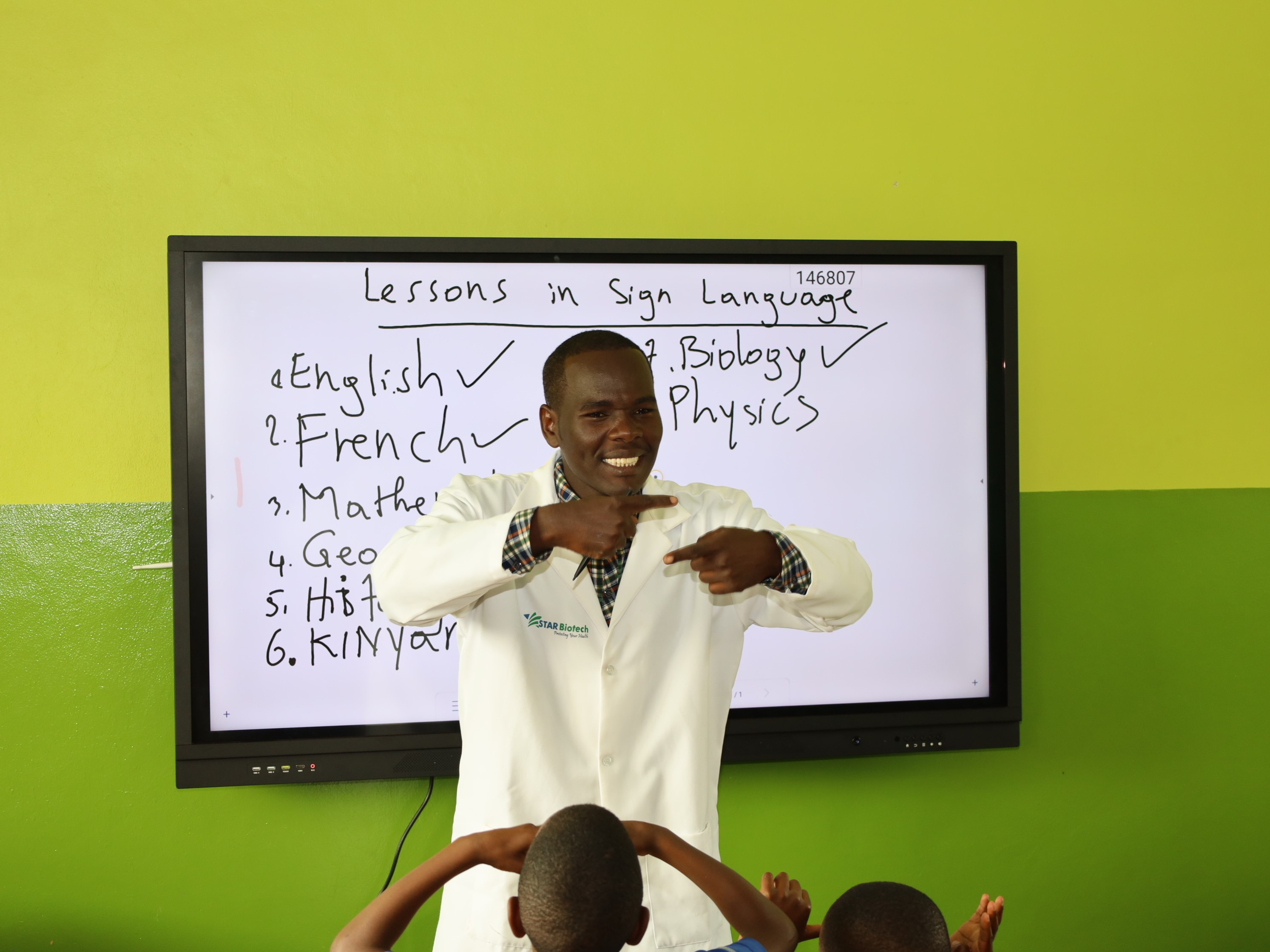 A teacher stands in front of the class, with a board behind saying 