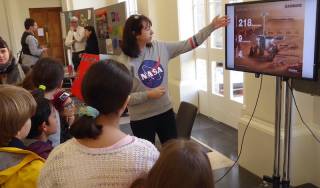 Female with NASA top talking to school pupils in front of a TV monitor showing a Mars Rover